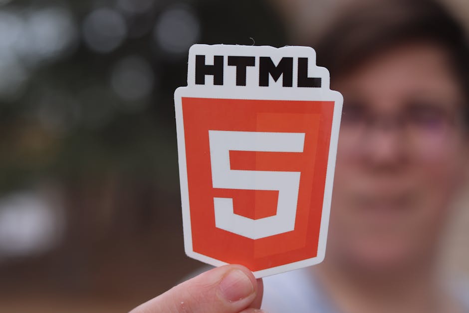 A person holds a sticker featuring the HTML5 logo, which consists of a stylized orange shield with a white number '5' in the center and the black text 'HTML' at the top. The background is blurred but 
