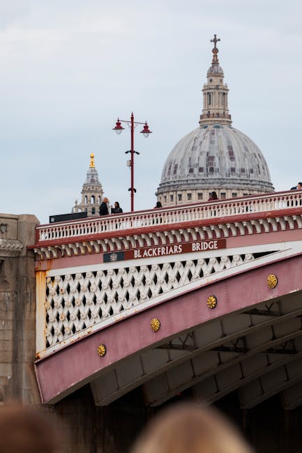 A wide view of the London skyline at sunset, featuring the Blackfriars Bridge crossing the River Thames in the foreground. The bridge has a detailed stone structure with decorative arches and street l