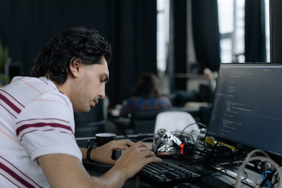 A young man with dark, wavy hair and light skin seated at a desk in an office environment, working on a black keyboard and looking closely at a large computer monitor displaying lines of code in a dar